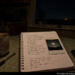 A kitchen table at night with a handwritten notebook, a blurred Polaroid of a luminous sky object, a coffee mug, and a single unexplained amber light visible through the window