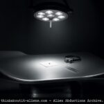 A tiny metallic alien implant rests on surgical gauze under harsh light on a contoured examination table, with the silhouette of a Grey alien barely visible in the surrounding darkness